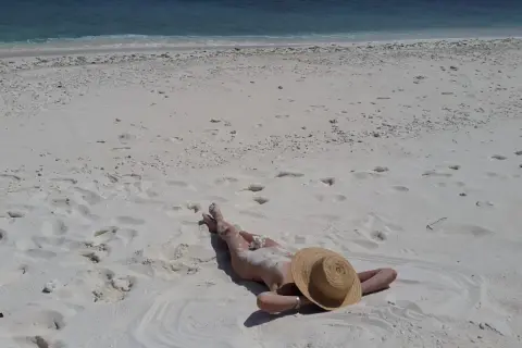 Person lying on a white sandy beach, wearing a straw hat, with the blue ocean in the background.
