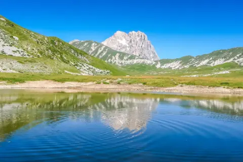 Specchio d'acqua riflette le maestose montagne del centro Italia, con verdi pendii e un picco roccioso sotto un cielo azzurro.