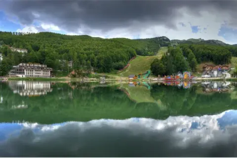 Lago alpino con riflessi di alberi e case, sullo sfondo il **passo del Cerreto** con piste da sci e giochi per bambini.