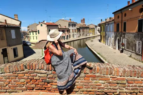 Donna con cappello di paglia e zaino rosso seduta su un ponte di mattoni a Comacchio, con canali e case colorate. Un luogo ideale per una gita con bambini.