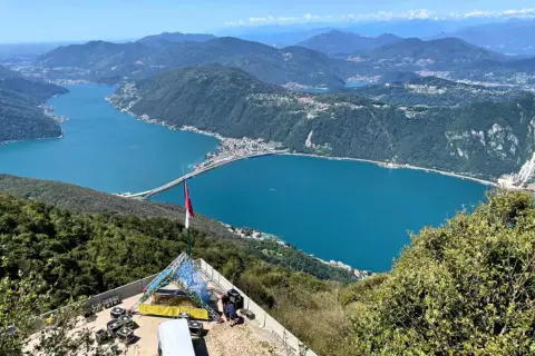 Vista panoramica del Lago di Lugano, con un ponte che lo attraversa. In primo piano, una struttura per eventi e una bandiera. Ideale per le val d'Intelvi passeggiate.
