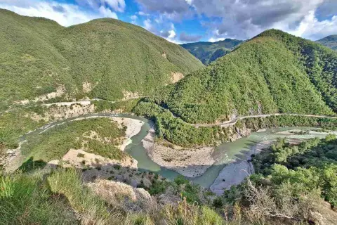 Un fiume serpeggia tra montagne verdi e scoscese, con una strada che costeggia il corso d'acqua. Il borgo di Brugnello si intravede tra la vegetazione lussureggiante.