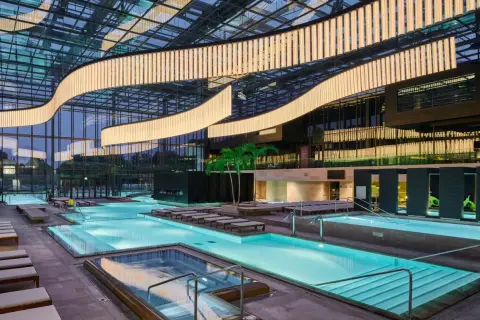 Interno moderno delle Merano Terme con piscine luminose, lettini e palme sotto un soffitto vetrato con illuminazione scenografica.