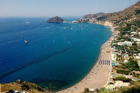 Vista aerea di una baia con spiaggia affollata, ombrelloni colorati e mare blu intenso. Un promontorio roccioso e un isolotto punteggiano l'orizzonte. Potrebbe essere una delle 5 spiagge più belle di Ischia.