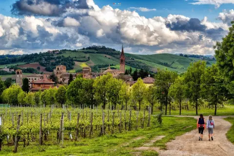 Due escursioniste passeggiano tra vigneti rigogliosi e dolci colline modenesi, con un borgo antico sullo sfondo.
