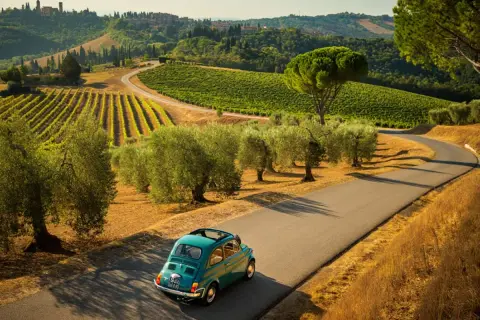 Un'iconica Fiat 500 azzurra percorre una strada tortuosa tra vigneti e ulivi nei **colli bolognesi**, con borghi arroccati all'orizzonte.