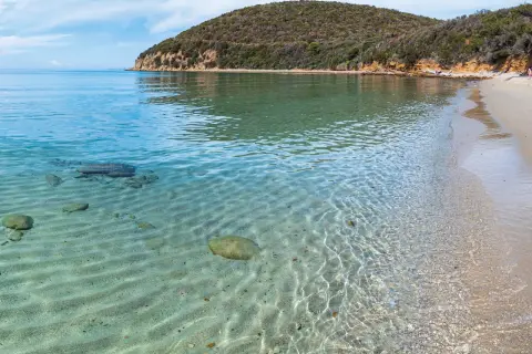 Acque cristalline e sabbia dorata nella Maremma Toscana. Il mare riflette il cielo azzurro, mentre la collina verdeggiante si affaccia sulla costa.