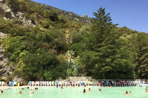 Piscina con persone che nuotano, circondata da vegetazione lussureggiante e rocce, un luogo che sembra uscito dalle grotte delle ninfe.