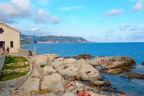 La chiesa bianca domina la costa rocciosa di Bordighera, con bagnanti che si godono il sole. Un luogo incantevole per scoprire cosa vedere a Bordighera.