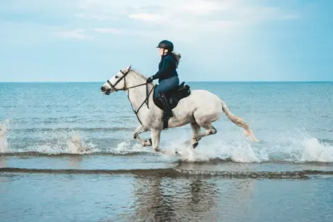 Una cavallerizza sperimenta una gioiosa passeggiata a cavallo sul mare, sollevando spruzzi d'acqua con il suo bianco destriero.