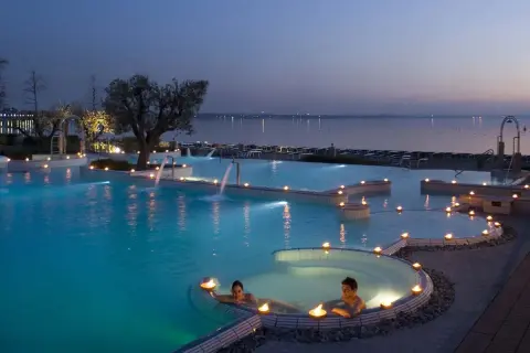 Coppia rilassata in una piscina termale illuminata da candele, con cascate e vista lago al tramonto. Un'oasi di pace alle terme a Brescia.