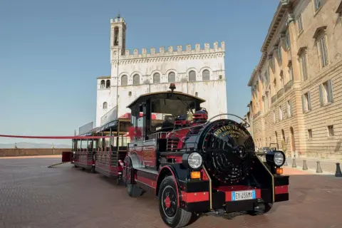 Il trenino turistico di Gubbio, rosso e nero, attende i passeggeri in una piazza storica con palazzi antichi.