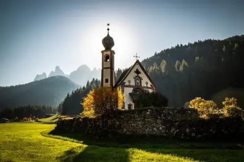 Chiesa di San Giovanni in Ranui, incastonata nel **Val di Funes**, con le Dolomiti sullo sfondo illuminate dal sole.