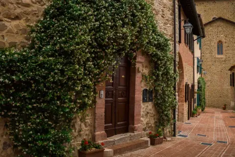 Un antico borgo in Umbria, con un portone di legno scuro avvolto da rampicanti fioriti. La strada in mattoni rossi sale dolcemente tra case in pietra.