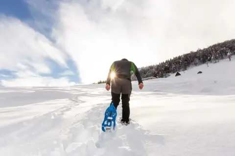 Escursionista con racchette da neve cammina su sentiero innevato, scoprendo posti belli in montagna Piemonte inverno. Sole splendente tra le nuvole.