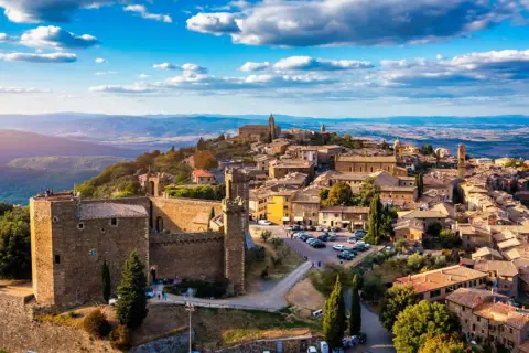 Scorci borghi antichi arroccati su una collina, con un castello imponente e case in pietra sotto un cielo azzurro con nuvole bianche.