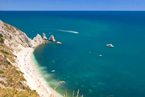 La spiaggia più bella del Conero, con sabbia bianca, mare cristallino e scogliere imponenti. Barche solcano le acque turchesi.