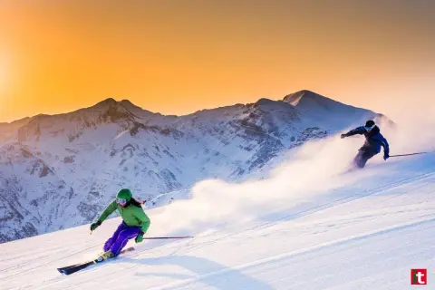Due sciatrici sfrecciano su un pendio innevato al tramonto, con montagne maestose sullo sfondo. Un'emozione da provare per chi cerca dove sciare in Emilia Romagna.