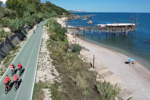 Ciclisti lungo la via verde della costa dei trabocchi, con il mare blu e le antiche macchine da pesca sullo sfondo.