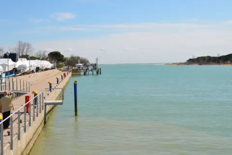 Un uomo guarda il mare calmo a Marano Lagunare, con barche coperte e un molo. La spiaggia lagunare è serena.