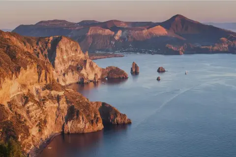 Scogliere dorate e mare blu intenso alle Isole Eolie, con piccole spiagge nascoste e faraglioni che emergono dall'acqua.