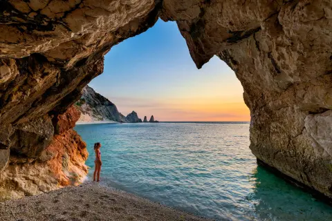 Vista da una grotta sulle spiagge della Riviera del Conero al tramonto. Una ragazza ammira il mare turchese e le rocce a picco.