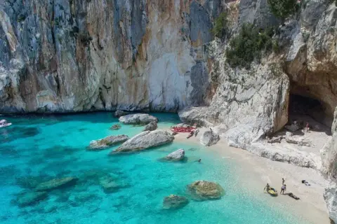 Spiaggia incantevole sulla costa di Baunei, con acque cristalline, rocce imponenti e una grotta marina. Kayak e persone godono del sole.