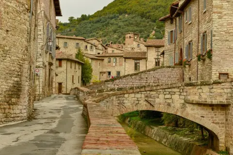 Antico ponte in pietra attraversa un ruscello, fiancheggiato da case in pietra e vicoli stretti, tipici dei paesi in Umbria.