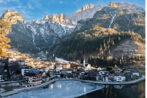 Villaggio alpino innevato ai piedi delle Dolomiti, ideale per escursioni invernali. Lago ghiacciato riflette le montagne.