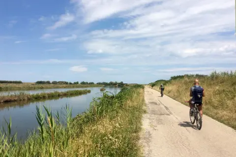 Cicloturismo in Emilia Romagna: due ciclisti pedalano su una strada sterrata lungo un canale, con vegetazione rigogliosa e cielo azzurro.