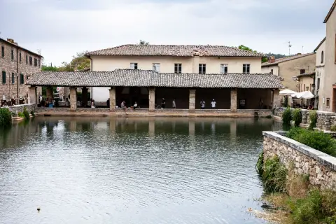 La piazza delle sorgenti termali di Bagni Vignone, con persone che si rilassano sotto un portico e il riflesso dell'acqua.