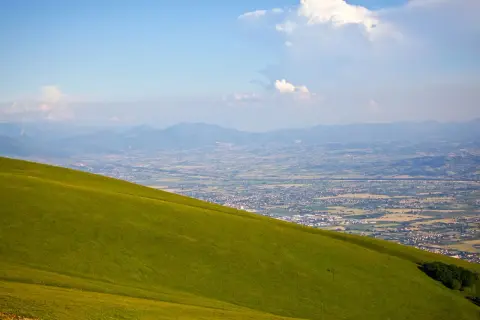 Prato verde scende dolcemente dal monte Subasio verso una valle punteggiata di case e montagne all'orizzonte.