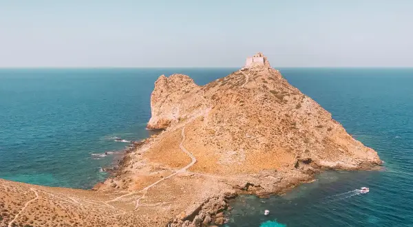 Rocciosa isola con un antico fortino in cima, circondata da acque turchesi. Un sentiero serpeggia verso la vetta, evocando le spiagge delle isole Egadi.