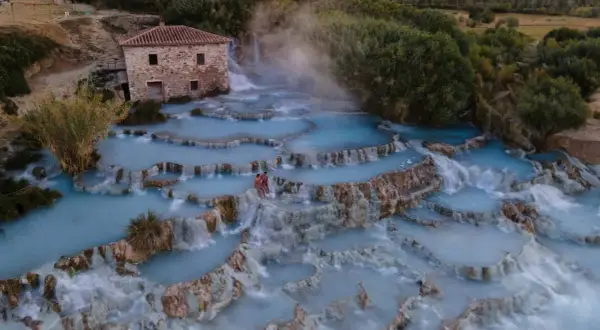 Cascate naturali con acqua azzurra e vapore, un casolare in pietra e due persone che si godono le terme di Saturnia.