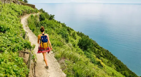 Donna con zaino cammina sul sentiero dell'Alta Via dei Monti Liguri, con il mare azzurro a destra e vigneti a sinistra.