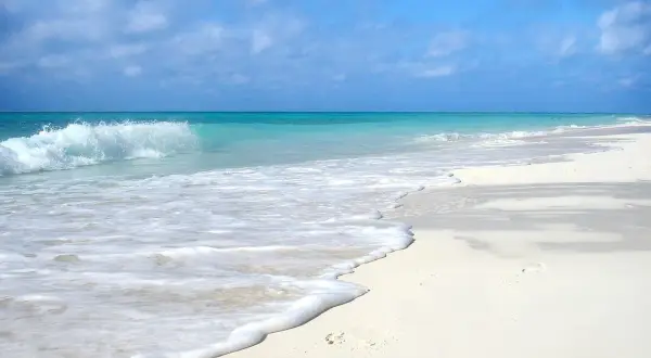 Onda che lambisce la sabbia bianca di una spiaggia d'inverno, con cielo azzurro e nuvole sparse.