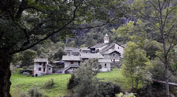Borgo in pietra con campanile, immerso nel verde della Val Chiuse lla. Un ponte attraversa il torrente.
