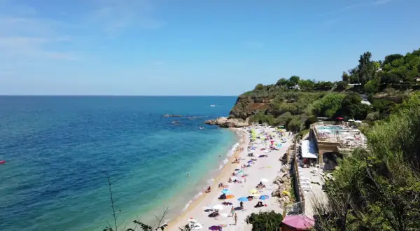 Spiaggia affollata con ombrelloni colorati, mare azzurro e scogliere verdi in Abruzzo. Un resort con piscina domina la scena.
