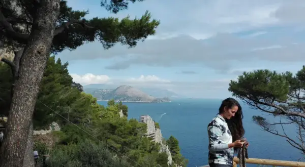 Donna con sciarpa guarda il cellulare, con vista mare e montagne di Capri a piedi.