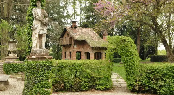 Un giardino incantato a Grazzano Visconti, dove si trova un'antica statua e una casetta in mattoni circondata da siepi verdi.