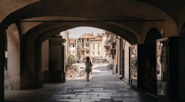 Una donna cammina sotto un portico verso una piazza soleggiata, con biciclette parcheggiate e palazzi storici che ricordano i paesi delle Langhe.
