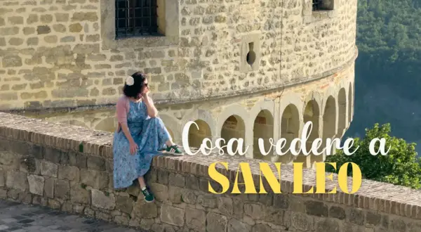 Donna seduta su un muretto di pietra a San Leo, con testo "Cosa vedere a San Leo".