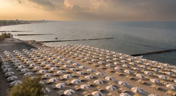 Filetto di sabbia dorata con file ordinate di ombrelloni e lettini, bagnato dal mare del Friuli Venezia Giulia al tramonto.