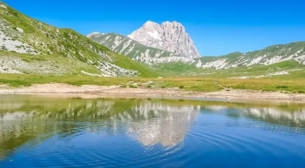Specchio d'acqua riflette le maestose montagne del centro Italia, con verdi pendii e un picco roccioso sotto un cielo azzurro.