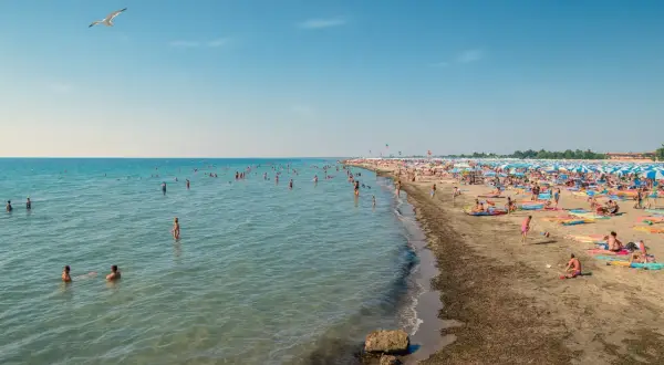 Gente in acqua e sulla sabbia, ombrelloni colorati e gabbiani in volo sulle spiagge friulane.