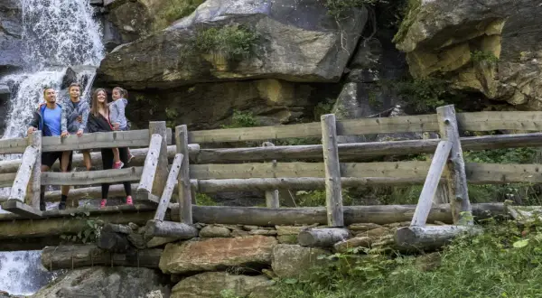 Famiglia felice sul sentiero delle cascate, con una cascata fragorosa alle spalle e un ponte di legno rustico.