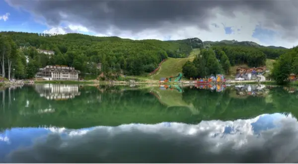 Lago alpino con riflessi di alberi e case, sullo sfondo il **passo del Cerreto** con piste da sci e giochi per bambini.