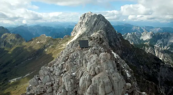 Bivacco Luca Vuerich, un cubo scuro su una cresta rocciosa, domina un panorama alpino mozzafiato sotto un cielo nuvoloso.