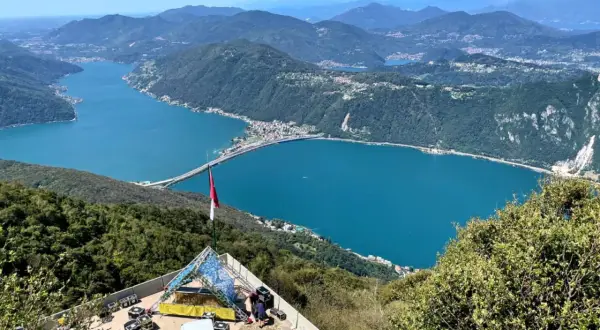 Vista panoramica del Lago di Lugano, con un ponte che lo attraversa. In primo piano, una struttura per eventi e una bandiera. Ideale per le val d'Intelvi passeggiate.