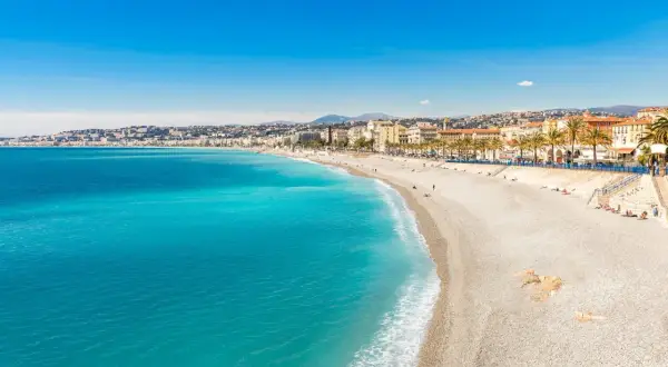 Spiaggia di ciottoli baciata dal sole sulla costa azzurra, con il mare turchese che lambisce la riva e palme che ombreggiano la Promenade des Anglais.